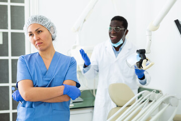 Upset asian woman in blue nurse uniform standing in dental clinic office on background with african american dentist expressing dissatisfaction. Concept of medical incompetence ..