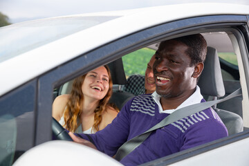 Portrait of laughing african american man driving car with cheerful fellow travelers in passenger seats. Friendly road trip and travel concept