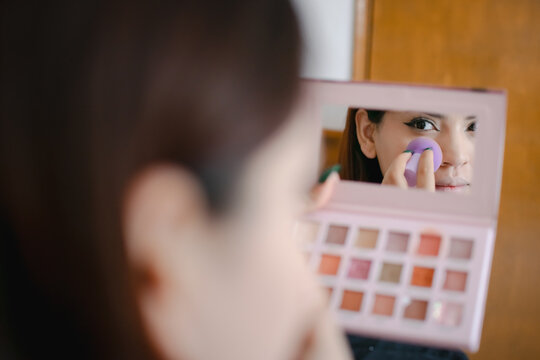 Primer plano en horizontal de una mujer joven latina aplic&aacute;ndose maquillaje por debajo de los ojos vi&eacute;ndose en un espejo peque&ntilde;o.