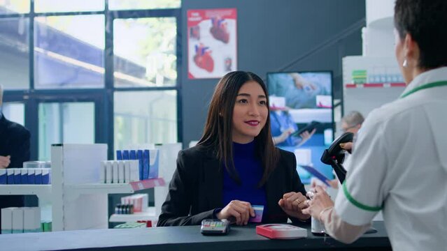 Asian woman in drugstore at checkout counter using credit card to purchase medical items. Happy customer doing contactless paying in pharmacy after finding needed virus stopping pills