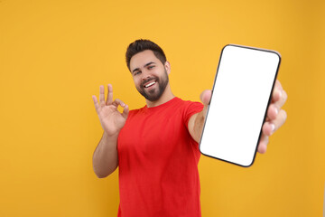 Young man showing smartphone in hand and OK gesture on yellow background