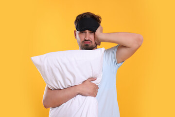 Unhappy man with pillow and sleep mask on yellow background. Insomnia problem