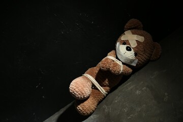 Stop child abuse. Tied toy bear with taped mouth and patches lying on grey floor against black...