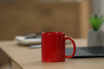 Red ceramic mug and laptop on wooden table at workplace