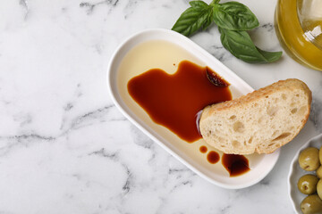 Bowl of organic balsamic vinegar with oil served with bread slice, basil and olives on white marble table, flat lay. Space for text