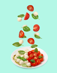 Fresh tomatoes. mozzarella cheese and basil leaves falling onto plate with Caprese salad against turquoise background