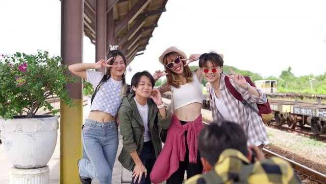 4K Group of Asian man and woman friends enjoy outdoor lifestyle using camera taking picture together during road trip low cost travel waiting for train at railway station on summer holiday vacation.