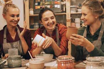 A company of three pretty young women friends make ceramic mugs in a pottery workshop. - obrazy, fototapety, plakaty