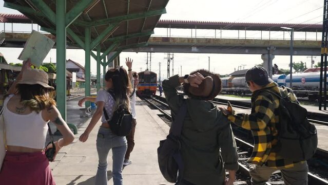 Group of Asian man and woman friends enjoy and fun outdoor lifestyle waiting for train at railway station during travel Thailand on summer holiday vacation. Youth culture and low cost travel concept.