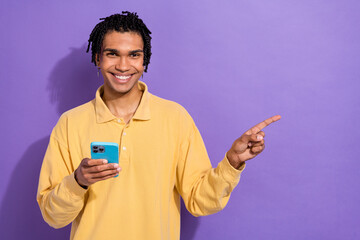 Photo of positive person with dreadlocks wear stylish shirt hold smartphone directing empty space isolated on purple color background