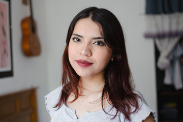 Mujer latina con cabello rojizo observando a la cámara con una sonrisa en un cuarto de fondo blanco y una guitarra en la pared