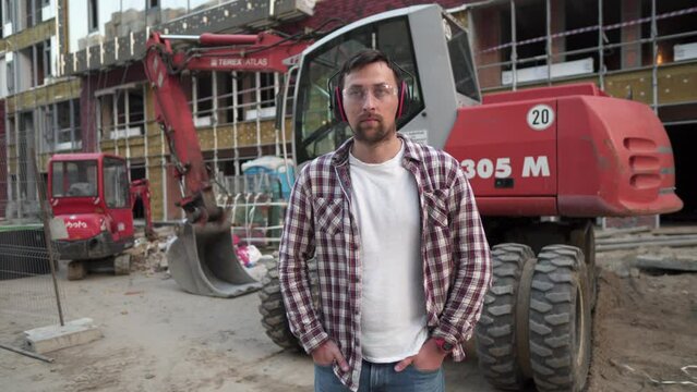 Portrait of successful builder, worker, contractor wearing hard hat and protective earphones, standing on work site with crossed arms confidently. Heavy construction equipment in background. 