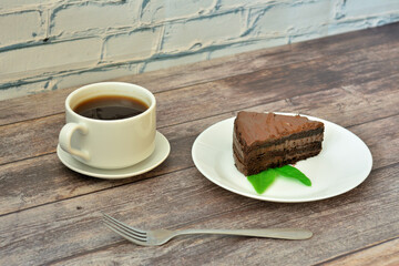 A plate with a piece of layered chocolate cake with mint leaves and a cup of hot coffee on a wooden table.