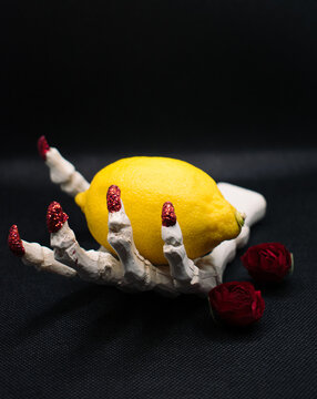 Skelleton hand with red nails holding yellow lemon on a black background with roses around