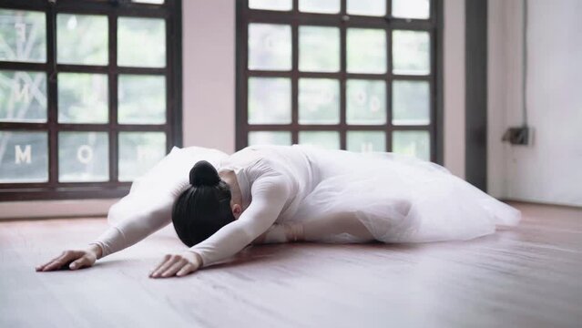 Young Asian ballet dancer in white tutu practicing ballet exercises bending forwards over her stretched body while sitting on the floor.