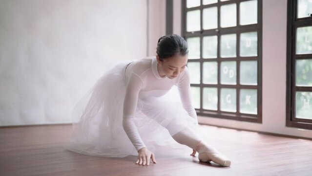 Young Asian ballet dancer in white tutu practicing ballet exercises bending forwards over her stretched body while sitting on the floor.