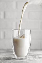 milk pouring in glass from jug on table with white brick wall as background