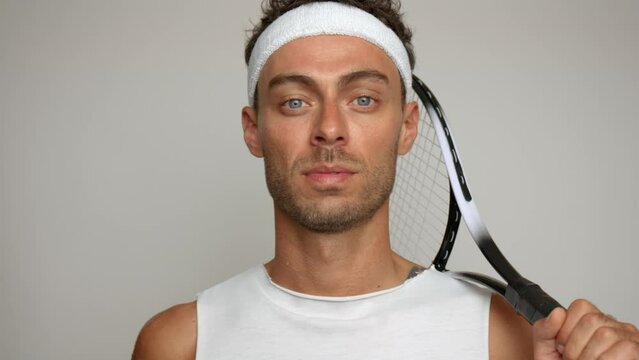 Handsome guy in white headband puts his tennis racket on shoulder, stands on white background, active lifestyle concept, copy space
