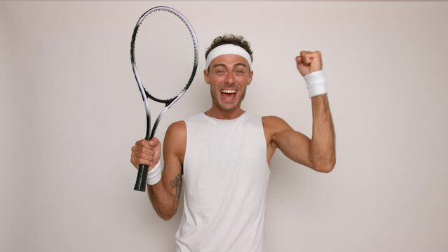 Young man won tennis set and raises racket in his fist up happily and smiles. wearing white top and white headband, active lifestyle concept, copy space