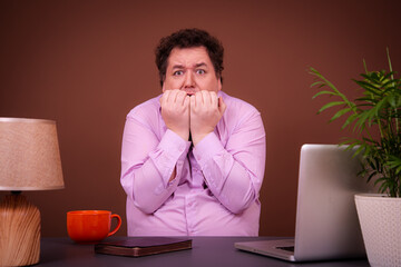 Funny fat man posing at a table with a laptop. Hard work online early morning Brown background.