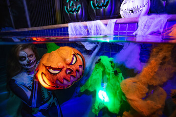 Halloween women holding a colourful pumpkin underwater in a pool with candys, skulls and spiders 