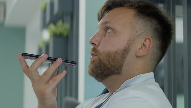 Male doctor sits in clinic cafe. Close up of professional medic talking by phone, recording voice message using social network or messaging app. Medical staff in hospital or medical center cafeteria.
