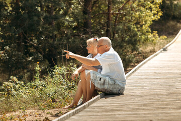 A happy elderly couple is sitting on a wooden path in a pine forest in the dunes and talking.