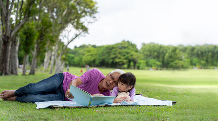 Happy Asian grandfather and granddaughter in the park spending free time together reading outdoors.