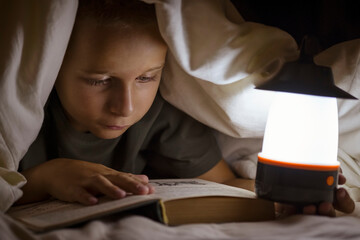Little boy is reading a book with flashlight under the blanket