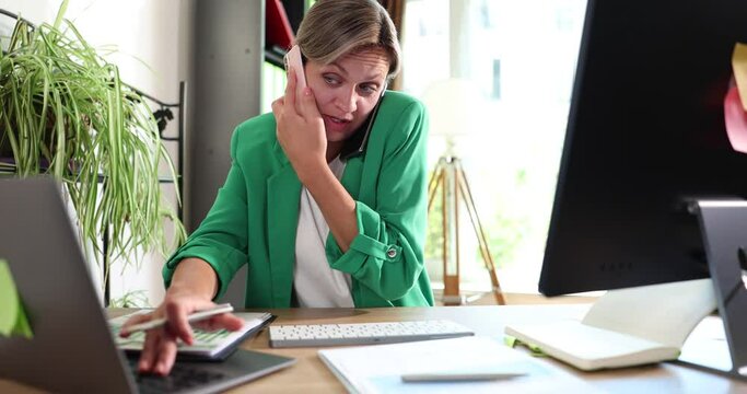Employee is multitasking working at computer in office and talking on phone. Manager looks at computer screen and consults with client on phone