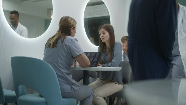 Female doctor sits in clinic cafe with patient. Professional medic discuss medical diagnostic results with woman. Digital tablet with MRI or CT brain scan image. Hospital or medical center cafeteria.