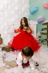 A little girl and a boy in red clothes have fun in the artificial snow by the fireplace in the studio. The girl is holding a box with gifts. Little kids in red christmas costume having fun beside a