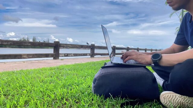 The teenage boy is working on his laptop computer on a green field, enjoying the clear weather and the happiness of working by the river.