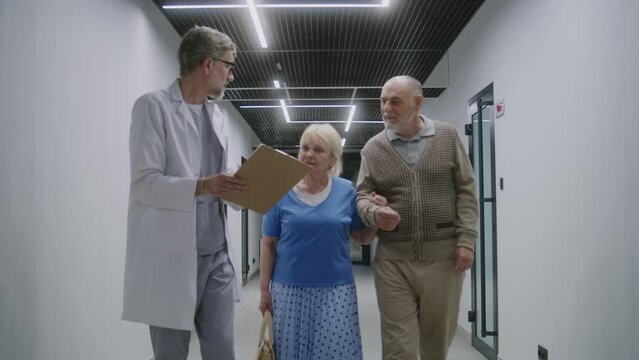 Elderly couple walks with doctor in clinic corridor. Doctor speaks with patients, discusses medical examination or test results. Medical staff and people in hospital or medical center. Healthcare.