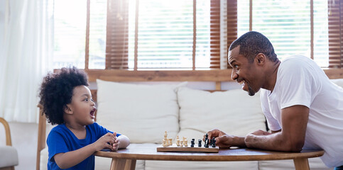 Portrait side view African black father little boy playing chess together, sitting at table, dad...