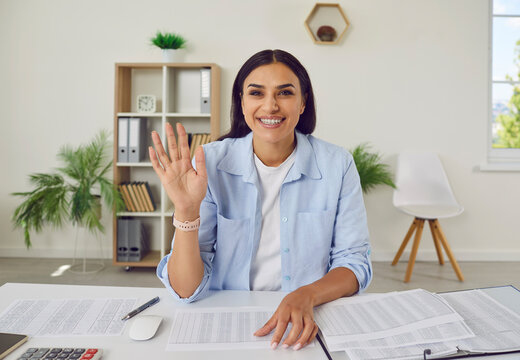 Portrait happy young woman business auditor accountant in casual shirt sitting at office desk with documents in front of camera, saying hello at online video call work meeting, waving hand and smiling