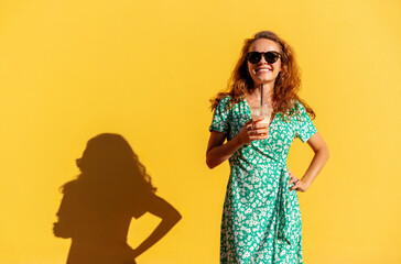 Young curly woman wearing trendy green dress and sunglasses holding glass of juice in hand and posing near yellow background.
