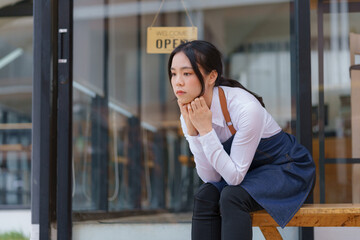 Unhappy and upset female entrepreneur sitting at her shop with an open signboard on the glass door.