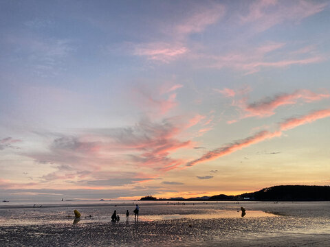 people standing on the beach at sunset.The sky is pink and orange, with clouds in the distance. The beach is sandy and there are some rocks in the foreground. The image is taken at sunset. South Korea