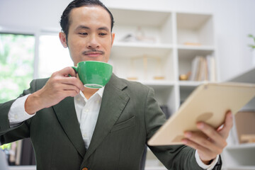 The manager of an Asian man sipping coffee on his desk in a radiant manner.
