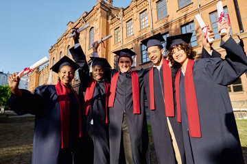 Group of happy students in gowns standing with diplomas outdoors near the university