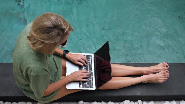 Female freelancer working with laptop in the swimming pool at sunny day. High quality 4k footage