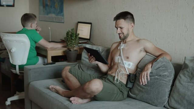 Man with heart sensors on body sits on couch and taps on tablet near child. Daily activities of father with 24-hour ECG monitoring medical device and son studying at laptop at table.