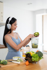 A slim Asian woman in gym clothes and headphones is making her healthy green smoothie.