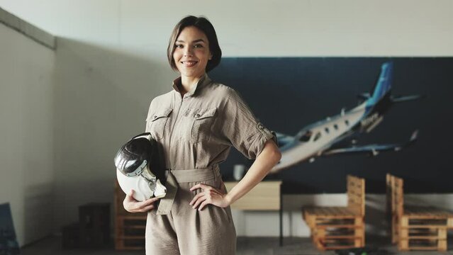 Good-looking young aviator turning head on camera after thoughtfully looking aside white standing in educational flying studio. Pretty lady in grey attire holding helmet in hand and gently smiling.