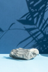 Empty podium made of gray stone on a blue background with a shadow of tropical leaves. Scene for the promotion of products, beauty, natural eco-cosmetics. Showcase, display case.