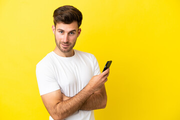 Young caucasian handsome man isolated on yellow background holding a mobile phone and with arms crossed