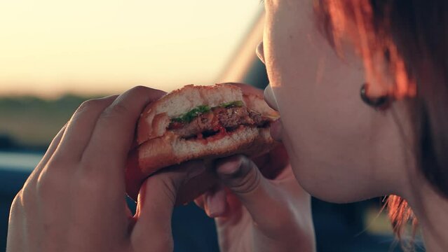 Woman eats tasty burger with patty sitting in car salon at sunset time