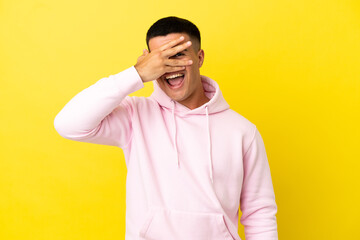 Young handsome man over isolated yellow background covering eyes by hands and smiling