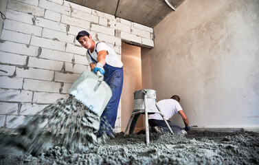 Male worker using shovel while shoveling sand-cement mix in building under construction. Man preparing floor screed material while standing near concrete screed mixer machine.